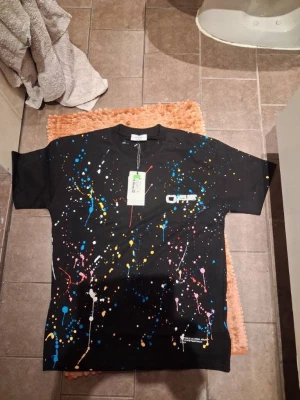 Off-White svart t-shirt med färgstänk - Svart t-shirt från Off-White med färgstänk i blått, gult, rosa och vitt. Tryckt Off-White-logga på bröstet och stort grafiskt tryck på ryggen. Klassisk passform och rund hals. Perfekt för dig som gillar streetwear och vill sticka ut.