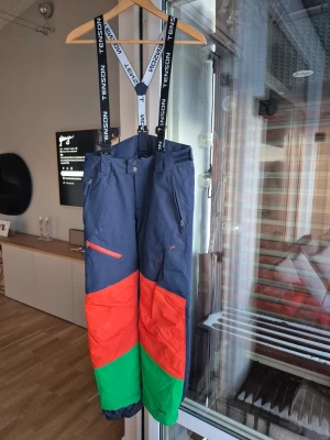 Skidbyxor från Tenson i färgblock - Feta täckbyxor från Tenson med cool färgblock-design i mörkblått, rött och grönt. Byxorna har justerbara hängslen med Tenson-logga, dragkedjor på fickorna och är perfekta för vinterns alla äventyr. Materialet är slitstarkt och vattenavvisande syntet.