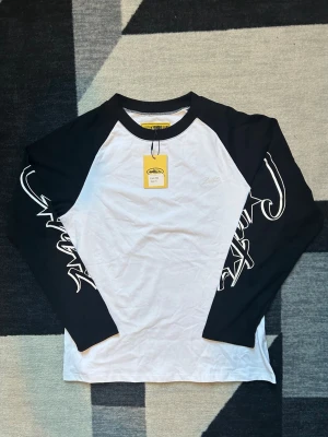 Svartvit långärmad topp Corteiz - Säljer en svartvit långärmad topp från Corteiz Guerillaz med kontrastfärgade ärmar och coolt tryck längs båda ärmarna. Toppen har rund halsringning och är gjord i 100% bomull för skön känsla. Perfekt för dig som gillar streetwear och grafiska detaljer.