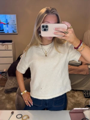Vit stickad kortärmad topp - Supermysig vit stickad topp med korta ärmar och rund hals. Modellen är lite croppad och har en enkel, stilren look som passar perfekt till jeans eller kjol. Materialet är mjukt och ger en chill vibe. Har en brun fläck 