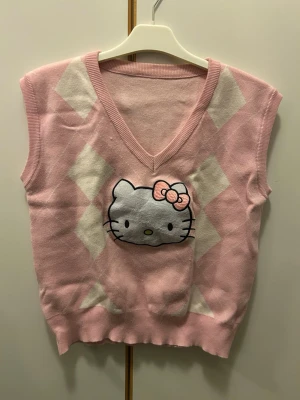 Hello Kitty rosa rutig stickad väst - Säljer en supersöt stickad väst i ljusrosa med vita rutor och ett stort Hello Kitty-tryck framtill. Västen har v-ringad hals och mjukt material som känns skönt mot huden. Perfekt för dig som gillar gulliga och lekfulla plagg med lite extra karaktär.