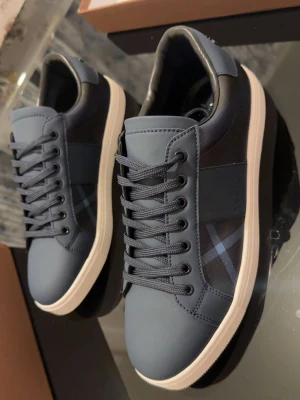 Blå sneakers från Burberry - Snygga blå sneakers från Burberry med klassiskt rutigt mönster på sidorna och vit platt sula. Skorna har snörning och detaljer i både skinn och textil. Burberry-logga bak på hälen. Perfekt för dig som gillar stilrena och exklusiva sneakers. Helt nya hör av dig vid intresse så hittar vi ett bra pris.