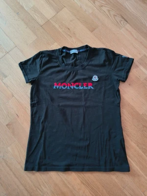 Svart Moncler t-shirt med paljetter - Svart t-shirt från Moncler med korta ärmar och rund hals. Framtill står Moncler i stora bokstäver med röda och blå paljetter. På bröstet sitter ett Moncler-märke och på ena ärmen finns en liten broderad detalj. Tillverkad i mjuk bomull.