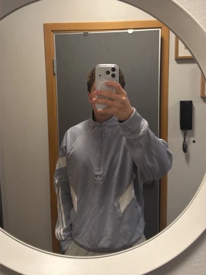 Ljusgrå/blå Adidas originals half zip sweatshirt - Säljer en ljusgrå Adidas sweatshirt med vita detaljer och klassiska tre ränder på ärmen. Tröjan har half zip med hög krage.