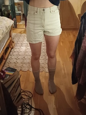 Högmidjade cream shorts - Säljer ett par ljusbeige högmidjade shorts i bomull med klassisk femficksmodell och uppvikta benslut. De har knapp och dragkedja framtill samt bälteshällor. Perfekta till sommaren och ger en clean och enkel look.