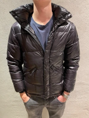 Replay jacka  - Säljer en svart pufferjacka med hög krage och dragkedja framtill. Jackan har två snedställda fickor med dragkedja och en glansig finish. Perfekt för kalla dagar och riktigt snygg till jeans eller sweatpants.