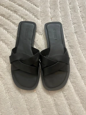 Svarta sandaler från Pull&Bear - Säljer ett par stilrena svarta sandaler från Pull&Bear Collection i storlek 37. De har breda korsade remmar och fyrkantig tå, vilket ger en modern look. Tillverkade i syntetmaterial med platt sula, perfekta för varma dagar.