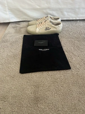 Saint Laurent vita sneakers i canvas - Säljer ett par klassiska vita Saint Laurent sneakers i canvas med snörning och rund tå. Skorna har Saint Laurent-logga broderad på sidan och detaljer i skinn. Snygg, minimalistisk design och medföljer original dustbag.