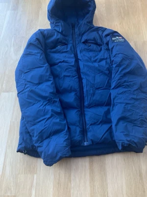 Blå pufferjacka från Sail racing - Säljer en blå pufferjacka från sail racing med huva och dragkedja framtill. Jackan har två bröstfickor med dragkedja och en patch med logga på ärmen.