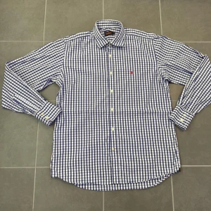Blårutig skjorta från Morris stl 41 - Säljer en klassisk blårutig skjorta från Morris i storlek 41. Skjortan har vit botten med blå rutor, knappar framtill och lång ärm. Broderad röd logga på bröstet och mjukt bomullstyg som känns skönt mot huden. Perfekt till jeans eller chinos.