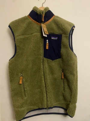 Grön fleeceväst från Patagonia - Säljer en helt ny mörk grön fleeceväst från Patagonia som var för liten i storleken för mig, den har marinblå detaljer.