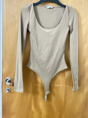 Beige långärmad bodysuit H&M - Säljer en beige långärmad bodysuit från H&M med rundad urringning och tryckknappar i grenen. Toppen har en tight passform och är tillverkad i ett stretchigt, mjukt material som sitter snyggt på kroppen.