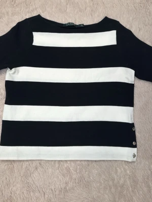 Randig topp från Ralph Lauren - Säljer en svartvit randig topp från Ralph Lauren med breda horisontella ränder. Toppen har rund hals, korta ärmar och tre dekorativa knappar i metall på sidan. Materialet är ribbat och känns mjukt och stretchigt.