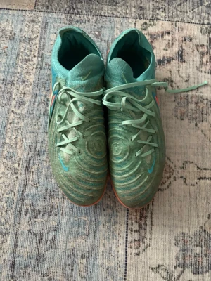 Nike turkosa fotbollsskor med snörning - Säljer ett par Nike fotbollsskor i turkos med coola cirkelmönster på ovansidan och en stor Swoosh-logga i orange och rött på sidan. Skorna har snörning och platt sula med dobbar i rosa och gult. Materialet är syntet och skon har en rund tå.