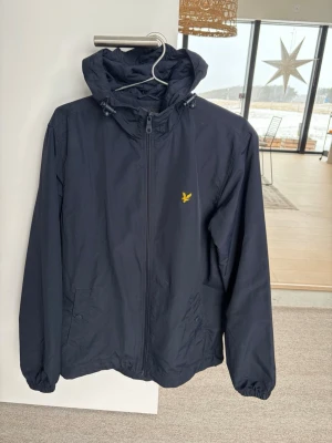 Mörkblå vindjacka från Lyle & Scott - Mörkblå vindjacka från Lyle & Scott med huva och gul broderad logga på bröstet. Jackan har dragkedja framtill, två fickor med knappstängning och elastiska muddar vid ärmslut. Perfekt för blåsiga dagar och enkel att matcha med olika outfits. Storlek medium 