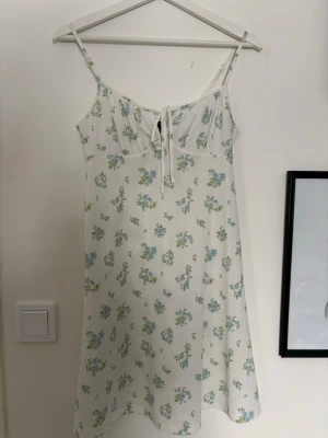 Blommig vit klänning från Gina Tricot - Supersöt vit kortklänning från Gina Tricot i storlek XS med blått och grönt blommönster. Klänningen har smala axelband, knytning fram och är tillverkad i ett lätt, strukturerat tyg. Perfekt för varma dagar och har en avslappnad passform.