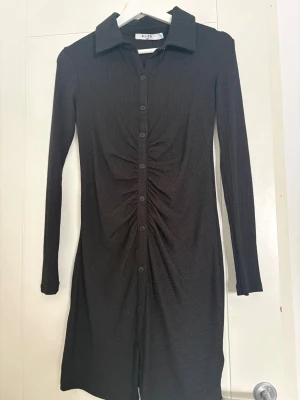 Svart ribbad skjortklänning NA-KD - Säljer en svart ribbad skjortklänning från NA-KD i storlek S. Klänningen har lång ärm, krage och knappar hela vägen framtill. Snygg rynkning längs knappraden ger en cool detalj. Materialet är mjukt och stretchigt, perfekt för dig som gillar stilrena plagg med twist.