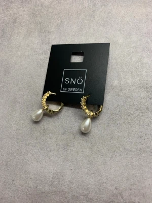 Guld pärlörhängen från Snö of Sweden - Säljer ett par snygga örhängen från Snö of Sweden med guldfärgade ringar och droppformade vita pärlor. Designen är elegant med små detaljer längs ringen som ger extra glans. Perfekt accessoar för att lyfta din outfit. Nypris på dessa är ca 800kr i butik. 