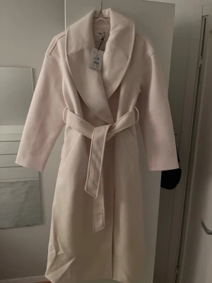Ljusbeige kappa från Gina Tricot (Helt oanvänd)  - Vit/cremefärgad lång kappa från Gina Tricot som är slutsåld överallt. Både knyte och knappar i midjan och i storlek Xs. Helt ny och oanvänd med prislappen kvar och utan defekter, säljer pga att den är för stor för mig. Skriv för frågor mm. (Den ser något rosa ut på bilderna vilket den inte är i verkligheten) 
