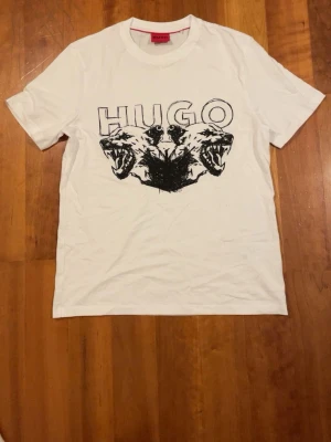 Vit t-shirt med tryck från Hugo - Vit t-shirt från Hugo med ett fett svart tryck på bröstet som visar två ritade varghuvuden och stor HUGO-logga ovanför. Klassisk rund hals och korta ärmar. Materialet är mjuk bomull och passformen är normal. Perfekt för dig som gillar grafiska prints och streetstyle.
