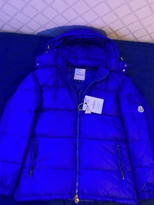 Blå Moncler pufferjacka med huva - Blå pufferjacka från Moncler med huva och två sidofickor med dragkedja. Jackan har klassisk Moncler-logga på ärmen och coolt serietryck på insidan. Fylld med dun och har en snygg, quiltad look. Perfekt för dig som vill ha både stil och värme under kalla dagar.