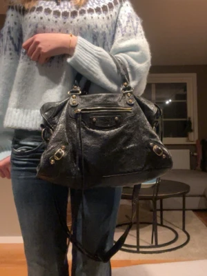 Svart Balenciaga handväska i skinn - Ny pris 27 000!! Säljer en Balenciaga i mjukt skinn med guldiga metalldetaljer och dragkedja i guld. Väskan har både handtag och axelrem, vilket gör den enkel att bära på olika sätt. Perfekt för dig som vill ha en klassisk och trendig accessoar. Har haft den länge så har tröttnat lite på den men är en väldigt snygg väska. Den är väldigt stor och rymlig. 