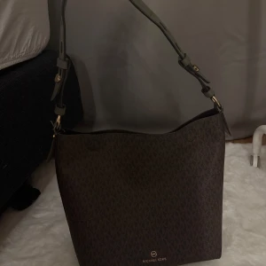 Brun axelväska från Michael Kors - Snygg brun axelväska från Michael Kors med MK-logga i guld på både remmen och väskans framsida. Väskan har ett diskret mönster med MK-bokstäver över hela ytan och justerbar axelrem i skinn. Perfekt storlek för att få plats med allt du behöver.