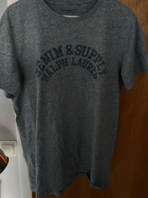Grå t-shirt från Ralph Lauren L - Säljer en grå t-shirt från Denim & Supply Ralph Lauren i storlek L. T-shirten har rund hals, korta ärmar och tryck framtill med märkesnamnet. Materialet känns mjukt och är troligtvis bomull, perfekt för chill dagar. Enkel och stilren design.