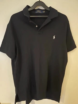 Svart piké från Polo Ralph Lauren - Klassisk svart pikétröja från Polo Ralph Lauren med vit broderad logga på bröstet. Tröjan har krage, korta ärmar och två knappar vid halsen. Tillverkad i mjuk bomull som är skön mot huden. Perfekt för en clean och stilren look.