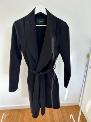 Svart kappa med bälte från VILA - Stilren svart kappa från VILA med krage och bälte i midjan. Jackan har lång ärm, två fickor fram och en klassisk omlott-design. Stl s.