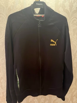 Svart ziptröja från Puma XL - Svart långärmad ziptröja från Puma i storlek XL. Tröjan har gul Puma-logga på bröstet, ribbad krage och dragkedja framtill. Fickor med dragkedja på sidorna. Perfekt för chill eller sportiga dagar.