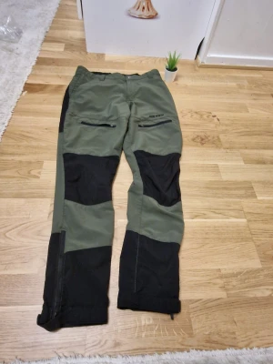 Gröna och svarta täckbyxor True North - Säljer ett par gröna och svarta täckbyxor från True North. Byxorna har dragkedjor på fickorna framtill, förstärkta partier på knän och bak, samt justerbara benslut med knapp och dragkedja. Perfekta för utomhusaktiviteter under kallare säsonger.