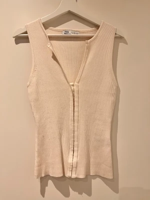 Popular linnetopp från Zara - Säljer en beige ribbad linnetopp från Zara i storlek L. Toppen är ärmlös med v-ringad hals och har en snygg knappdetalj framtill. Materialet är mjukt och stretchigt, perfekt för varma dagar. Enkel och stilren design som funkar till många olika looks.