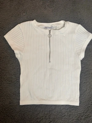 Vit ribbad topp med dragkedja från Zara - Säljer en vit ribbad topp från Zara med korta ärmar och rund hals. Toppen har en snygg silverfärgad dragkedja framtill med ringdetalj. Materialet är mjukt och stretchigt, perfekt för en chill och stilren look.
