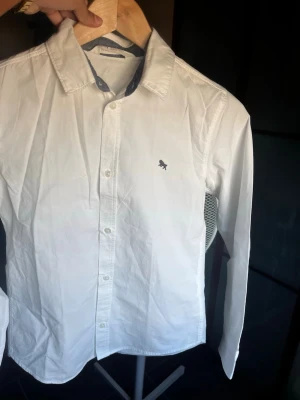 Vit skjorta från H&M med brodyr - Vit skjorta från H&M i bomull med klassisk krage och blå detaljer på insidan av kragen. Skjortan har knäppning framtill och en liten broderad hund på bröstet. Perfekt för en clean och stilren look.