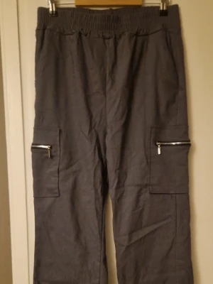 Grå cargopants med dragkedjor - Säljer ett par grå cargopants i storlek XL med elastisk midja och praktiska fickor med dragkedjor på sidorna. Byxorna är tillverkade av 95% polyester och 5% spandex vilket gör dem både bekväma och flexibla. Perfekt för dig som gillar streetwear och funktionella detaljer.