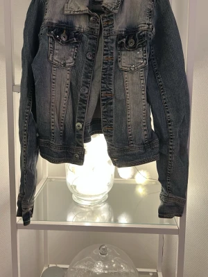 Mörkblå jeansjacka från Lindex - Snygg mörkblå jeansjacka från Lindex med klassisk krage, knappar framtill och två bröstfickor med lock. Jackan har lång ärm och cool tvättad look som ger en avslappnad vibe. Perfekt att slänga på över en hoodie eller t-shirt.