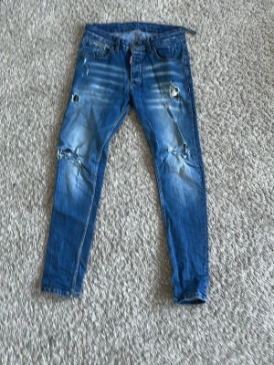 Blå ripped jeans från Dsquared2 - Säljer ett par blå jeans från Dsquared2 med snygga slitningar och hål på knäna. Jeansen har en smal passform och klassisk femficksdesign. Materialet är jeans med coola tvättade detaljer och Dsquared2-logga på knapp och etikett.
