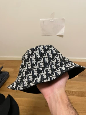 DI0R Hat - Säljer en bucket hat med ett snyggt rutigt mönster i blått och vitt. Hatten har en klassisk form med bred brätte och är tillverkad i ett grovt tyg som ger en cool och trendig look. Perfekt accessoar för att lyfta din outfit.