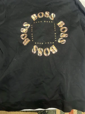 Svart sweatshirt från Hugo Boss - Säljer en svart sweatshirt från Hugo Boss med ett stort cirkulärt BOSS-tryck i guld och silver på bröstet. Tröjan har rund hals och är långärmad, perfekt för dig som gillar stilrena och lyxiga märkesplagg.