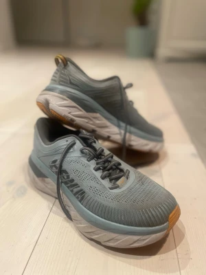 Hoka One One Bondi 7 sneakers grå - Säljer ett par Hoka One One Bondi 7 sneakers i grått med ljusblå och orange detaljer. Skorna har en tjock, dämpad sula och ventilerande mesh-överdel. Perfekta för dig som gillar att gå långt eller springa. Klassisk snörning och rund tå.