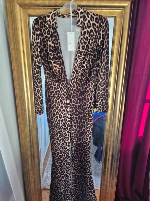 Leopardmönstrad byxdress med knappar - Snygg byxdress med leopardmönster, djup v-ringning och långa ärmar. Byxdressen har krage och knappar framtill, samt figurnära passform och vida ben. Perfekt för dig som vill sticka ut med ett djuriskt print.