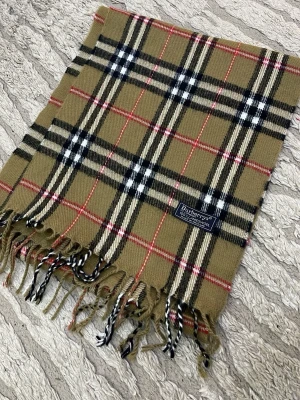 Burberry halsduk beige - Säljer en klassisk väst från Burberrys i beige med rutigt mönster i svart, vitt och rött. Västen är tillverkad i 100% lammull och har fransar längs nederkanten. Perfekt för dig som gillar tidlös design och vill ha något som sticker ut.