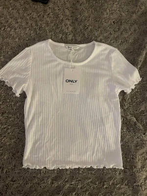 Vit ribbad topp från ONLY XS - Säljer en fräsch vit ribbad topp från ONLY i storlek XS. Toppen har korta ärmar, rund halsringning och vågiga kanter vid ärmslut och nederkant. Perfekt för dig som gillar enkel och stilren design med lite extra detalj på kanterna.