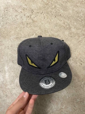 Grå Fendi keps med gula ögon - Säljer en grå keps från Fendi med coola gula ögon broderade framtill. Kepsen har en klassisk snapback och Fendi-logga på insidan och baksidan. Materialet är mjukt och känns som bomull, perfekt för att lyfta din streetstyle.