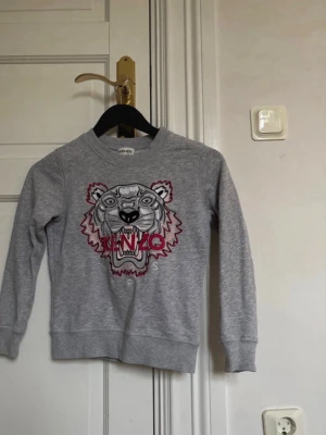 Grå Kenzo sweatshirt med tigertryck - Säljer en grå sweatshirt från Kenzo Kids med ikoniskt tigerbroderi och texten 'KENZO PARIS' framtill. Tröjan har rund hals, långa ärmar och ribbade muddar. Storlek 140, 10 yrs.