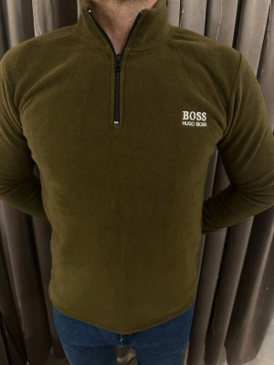 Olivgrön fleece från Hugo Boss - Säljer en olivgrön fleece från Hugo Boss med broderad logga på bröstet. Tröjan har hög krage, lång ärm och en praktisk half zip-dragkedja framtill. Perfekt för kyliga dagar och enkel att matcha med jeans eller joggers.