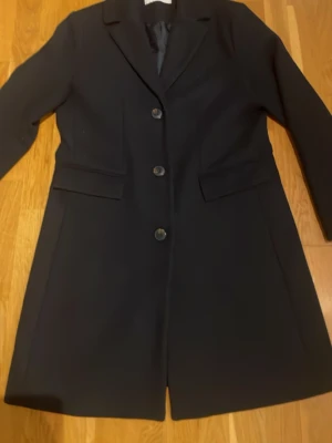 Mörkblå kappa med knappar - Säljer en stilren mörkblå kappa från Ellos med klassisk krage och tre knappar framtill. Jackan har två fickor med lock och är långärmad. Perfekt för dig som gillar en clean och tidlös look.