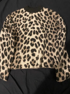 Leopardmönstrad stickad tröja - Säljer en stickad tröja med leopardmönster i beige och svart. Tröjan har rund hals med ribbad kant och långa ärmar. Perfekt för dig som vill sticka ut med ett djuriskt mönster och samtidigt hålla dig varm under kyliga dagar.