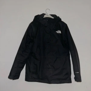 Svart vindjacka från The North Face - Svart vindjacka från The North Face med huva och vit logga på bröstet. Jackan har dragkedja framtill, två sidofickor med dragkedja men den ena går ej att stänga och justerbara muddar. Materialet är slitstark polyester och jackan har en clean, sportig look.
